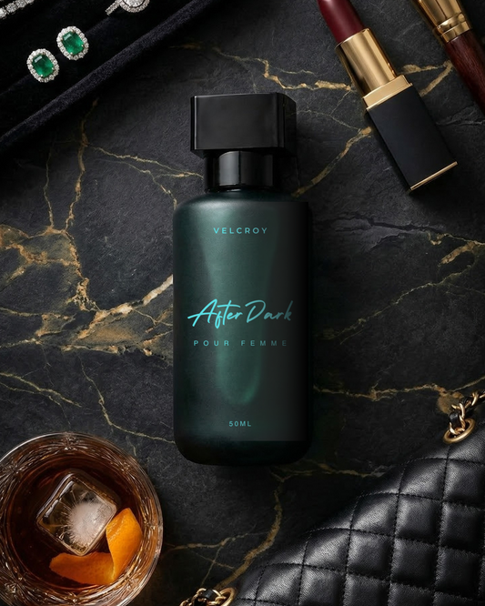 Perfume bottle labeled 'After Dark' surrounded by jewelry, lipstick, and a cocktail on a dark surface.