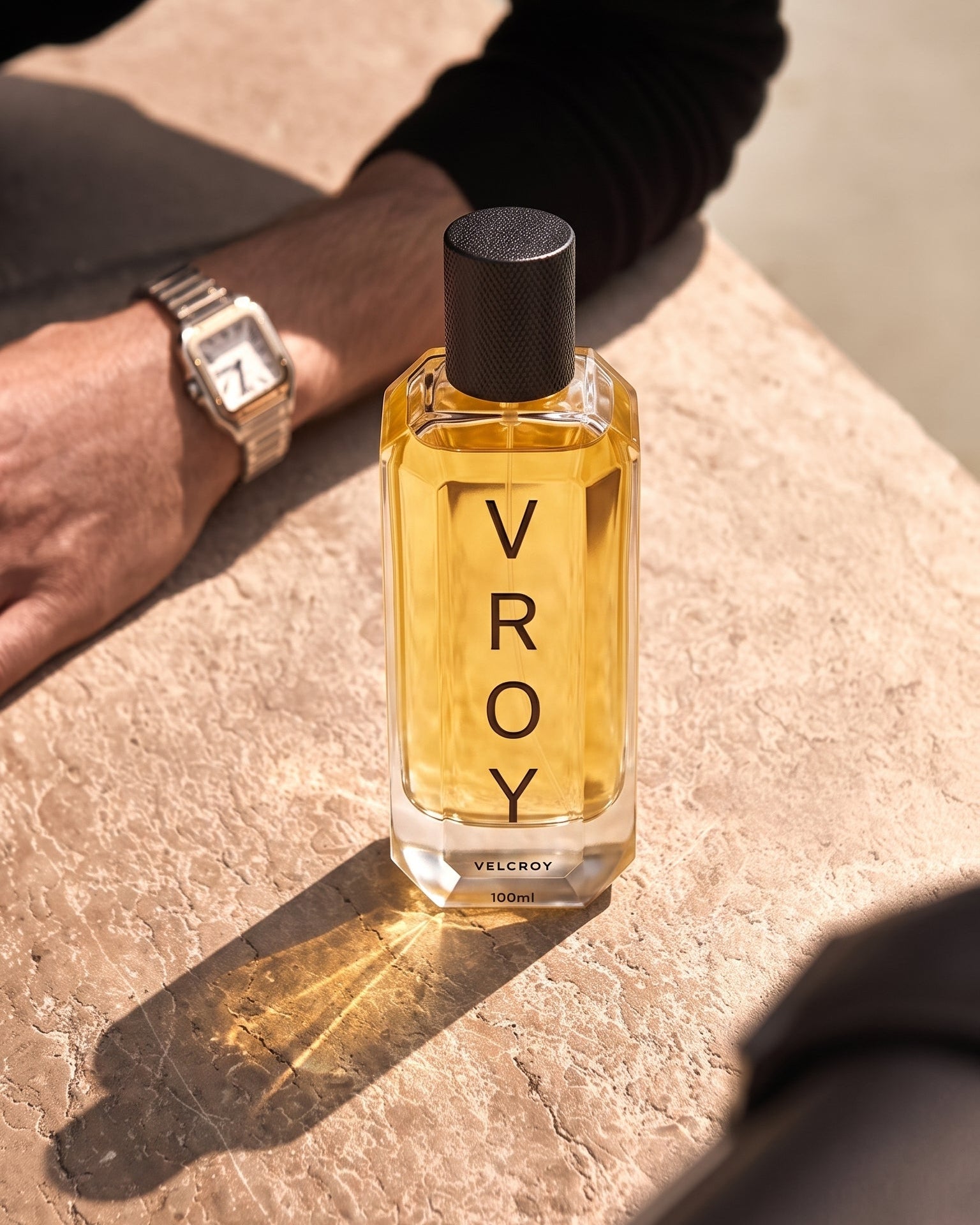 A clear glass bottle of VROY perfume with amber-yellow liquid and a black cap, standing on a textured stone surface outdoors. A person's arm and hand wearing a silver watch are visible in the background.